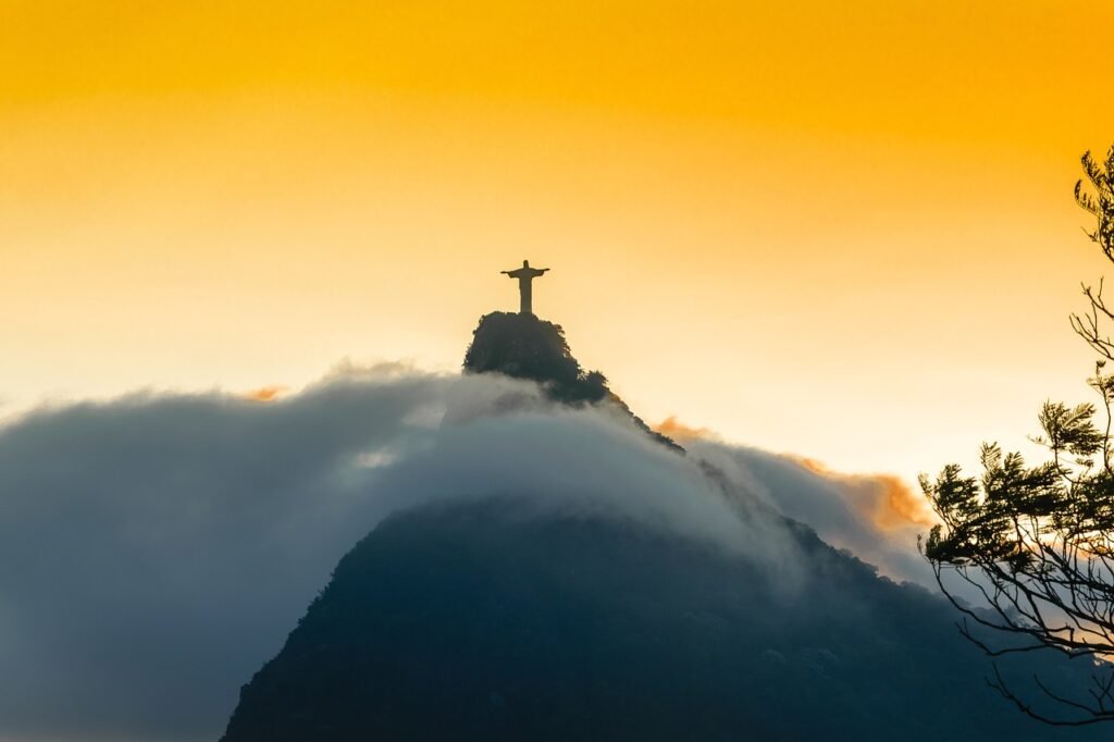 Cristo Redentor: como chegar, ingressos e horários 5 Foto do Cristo Redentor Tirada de longe. Nuvens ao redor e por do sol amarelado ao fundo.