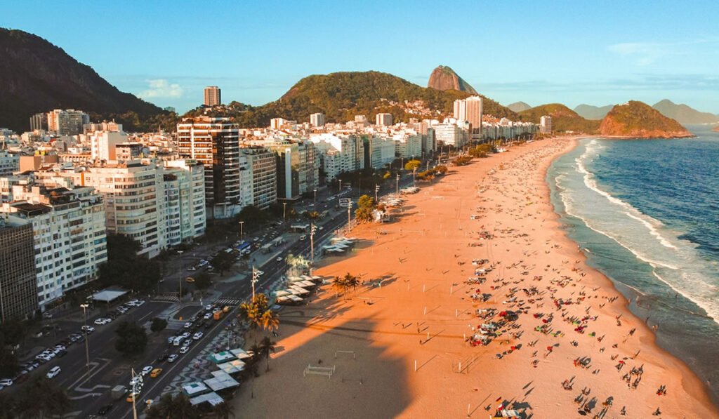 O que fazer no Rio de Janeiro? 10 pontos turísticos 1 Foto em dia de sol da areia e mar da praia de Copacabana, Rio de Janeiro; Pessoas na areia.